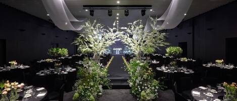 Un salón de banquetes con mesas dispuestas para un evento formal, con un arco floral central y un techo moderno oscuro con iluminación empotrada.