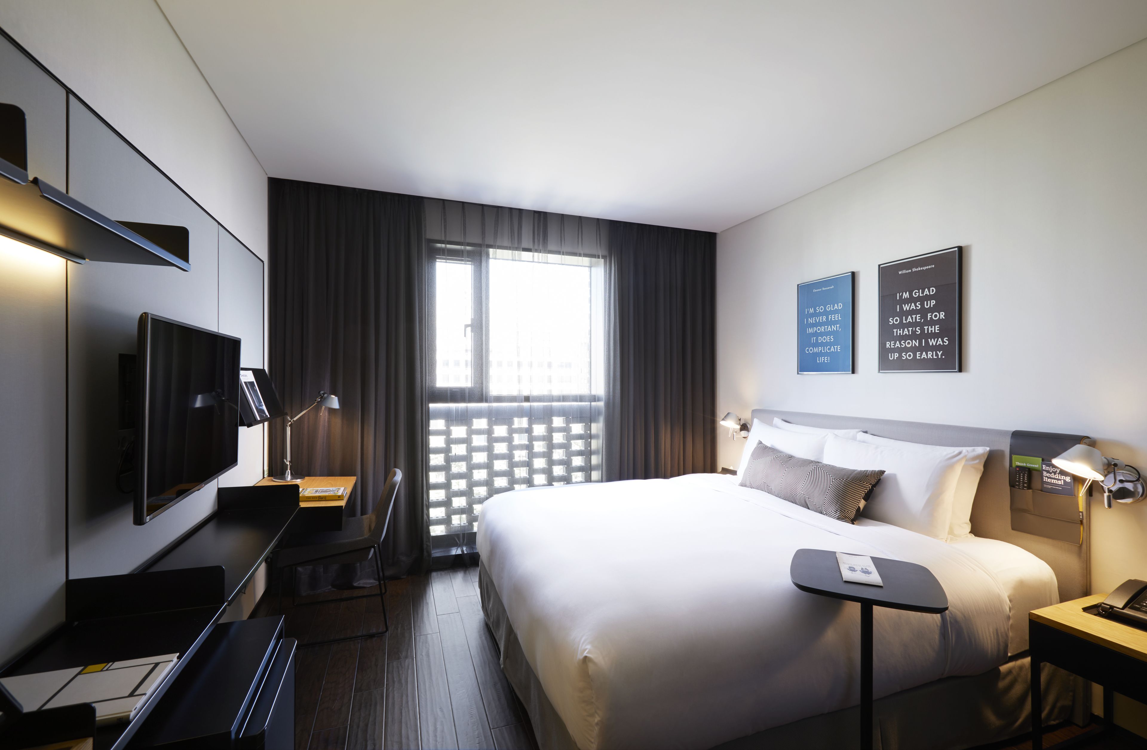 A modern hotel room with a large bed, a desk, a flat-screen TV, a desk lamp, and two motivational posters on the wall.