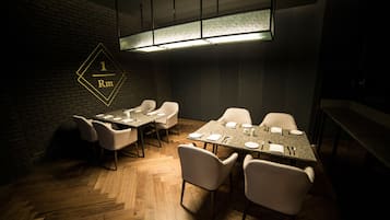 Un salón de conferencias con suelo de madera en patrón de espiga, sillas blancas, una larga mesa y una pared de ladrillo.
