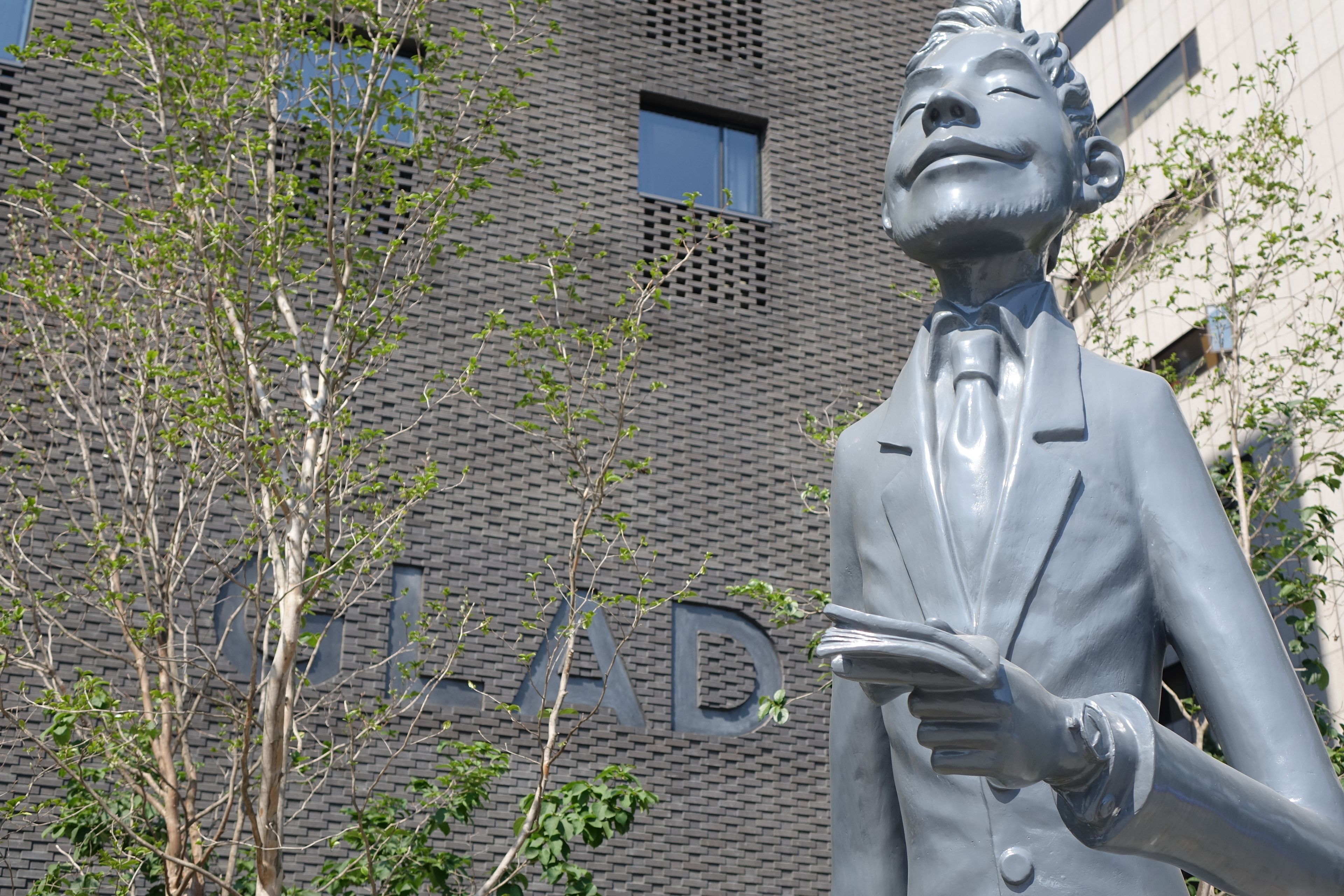 Una estatua de un hombre con traje sosteniendo un periódico, con un edificio al fondo.