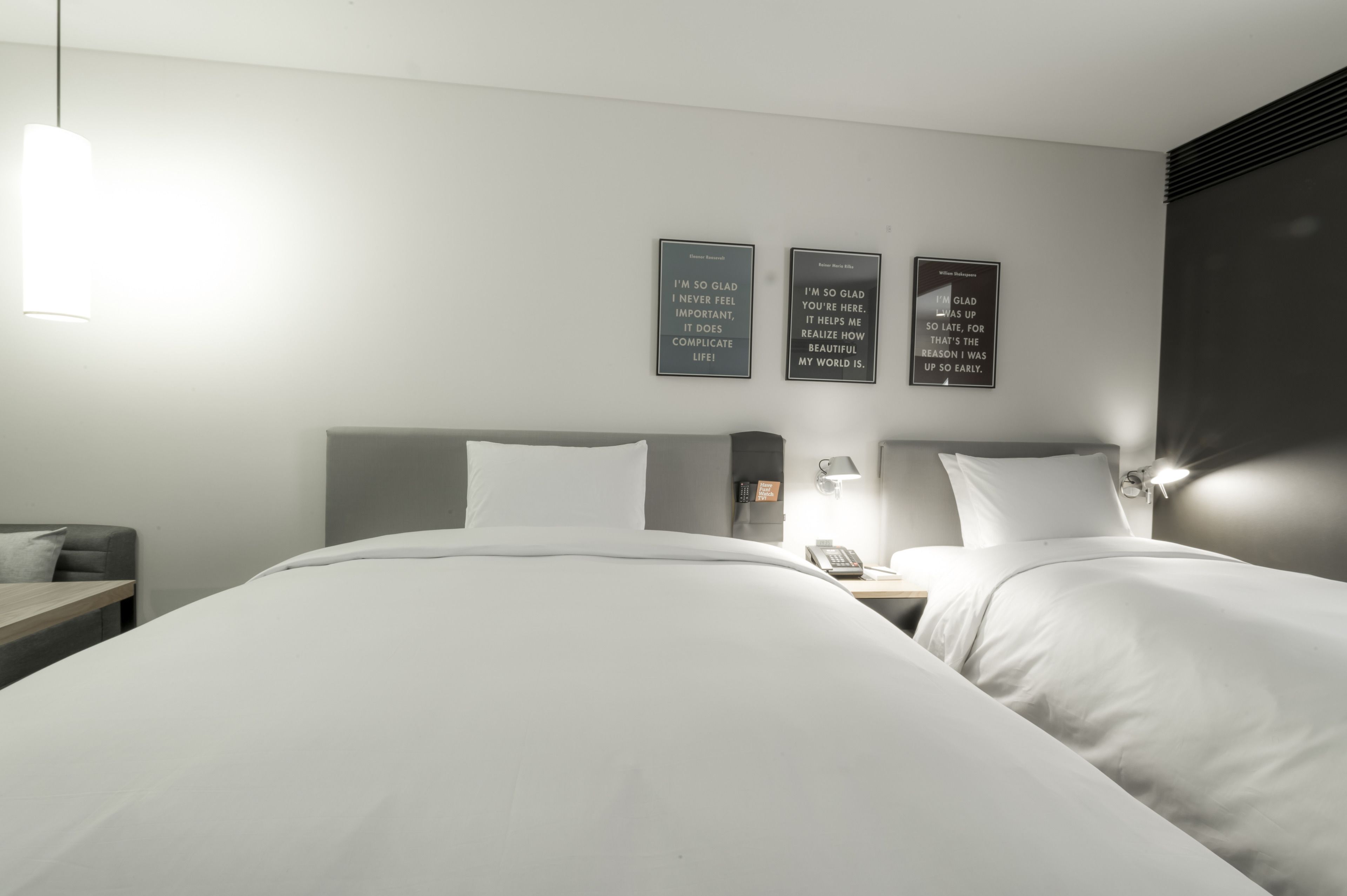 A modern hotel room with two beds, a bedside table with a lamp, and three motivational wall art pieces.