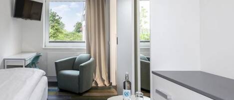 Una habitación de hotel moderna con una cama, un sillón gris, una mesita con una botella y un televisor montado en la pared.