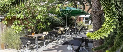 Une terrasse extérieure avec des tables et des chaises, entourée de verdure et d’un palmier.