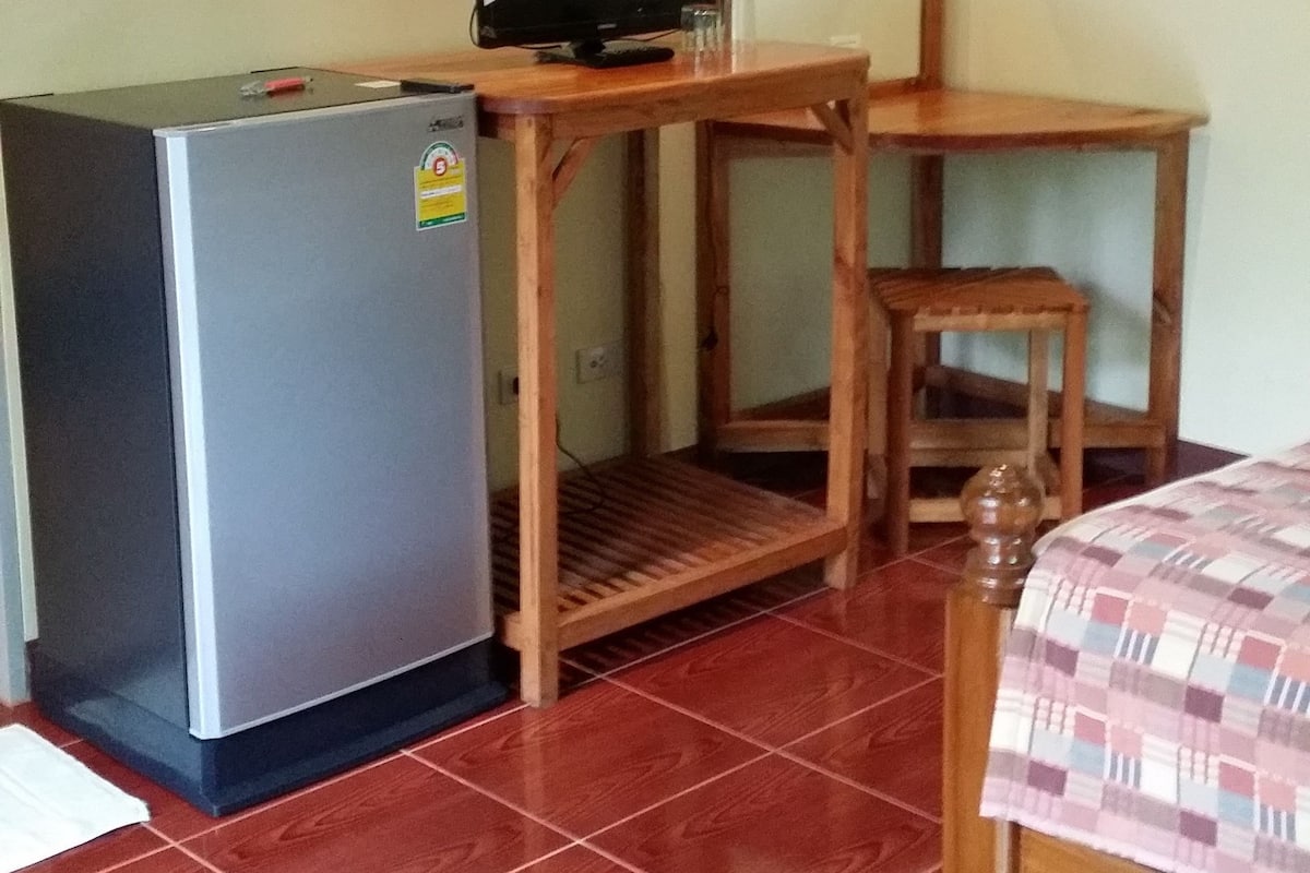 Une chambre de taille modeste, équipée d’un réfrigérateur, d’un bureau en bois avec une chaise et d’un lit recouvert d’une couverture à carreaux.