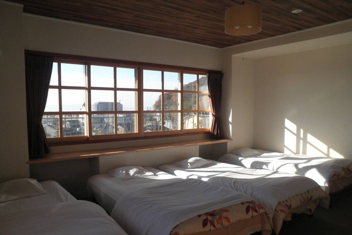A room with three beds, a large window with curtains, and a wooden ceiling.