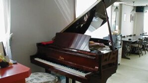 A grand piano in a room with a dining area and a window with curtains. - Gamagori Orange Pension (Gamagori)