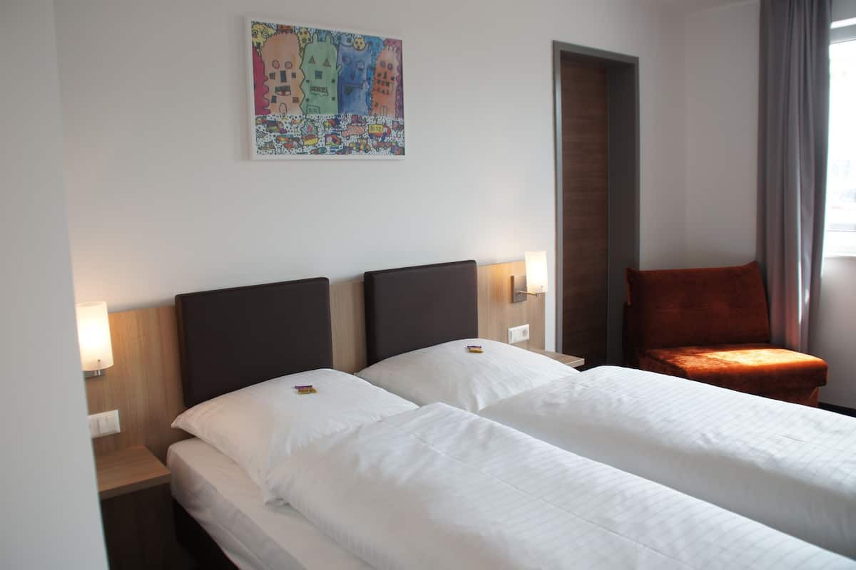 Habitación de hotel con dos camas, una mesita de noche con lámpara, una silla y un cuadro de colores en la pared.