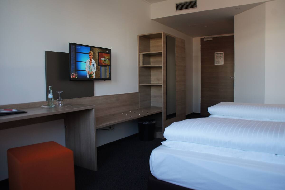 Una habitación de hotel con una cama, un escritorio con una computadora, un televisor y un taburete pequeño color naranja.