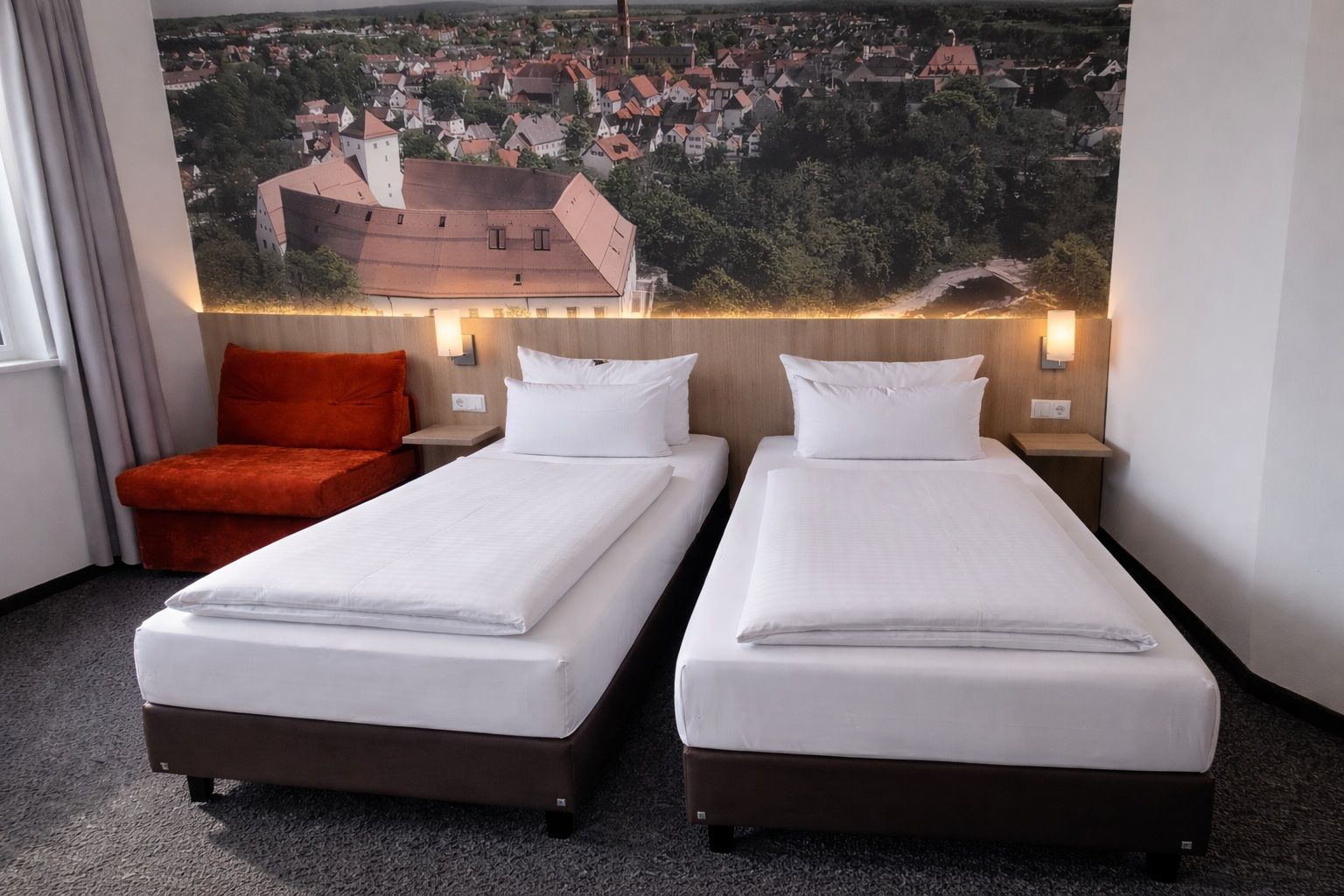 A hotel room with two single beds, a red chair, and a large cityscape painting on the wall.