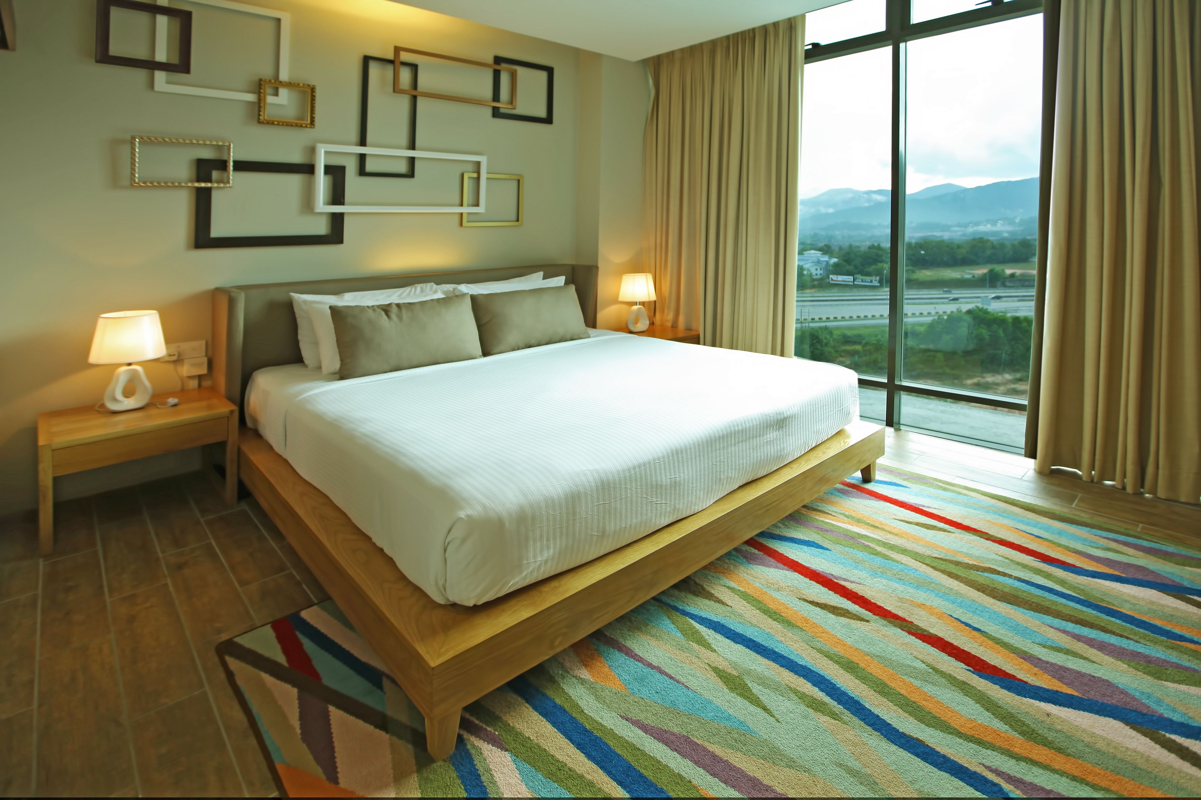 A hotel room with a large bed, two bedside tables with lamps, a colorful striped rug, and a view of a landscape through large windows.