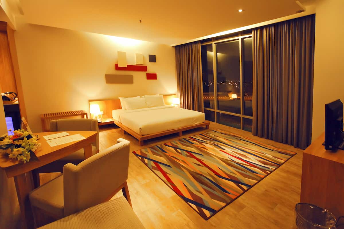 Habitación de hotel con una cama grande, un escritorio con una silla, una lámpara de escritorio y vistas a la ciudad de noche.