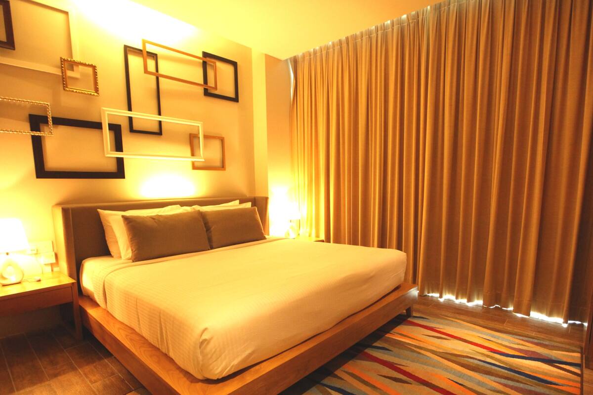 Habitación de hotel con una cama grande, lámparas de noche, una mesita de noche de madera, una alfombra colorida a rayas y una cortina con líneas verticales.