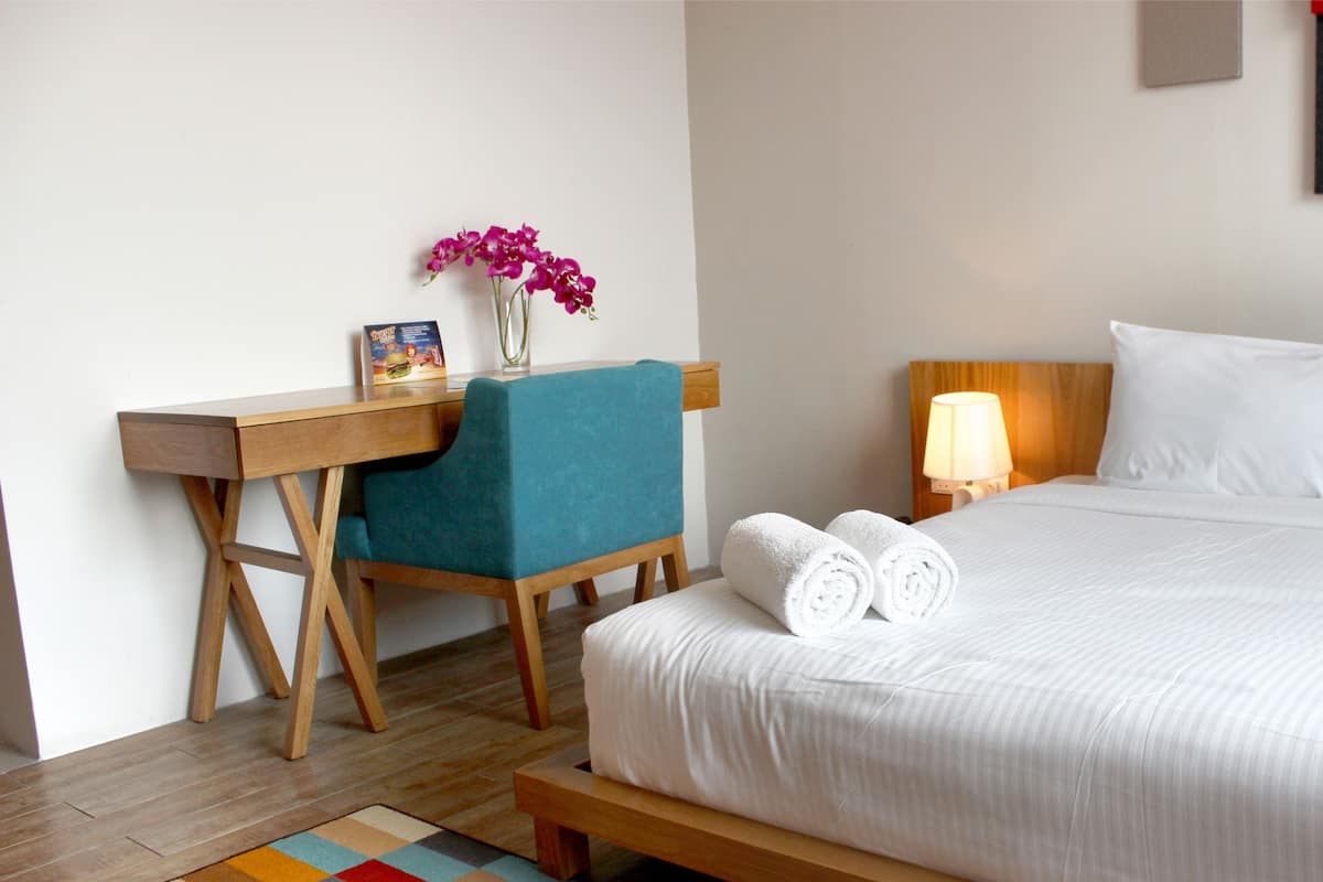 Habitación de hotel con cama, escritorio, silla, lámpara y una mesita con un florero.