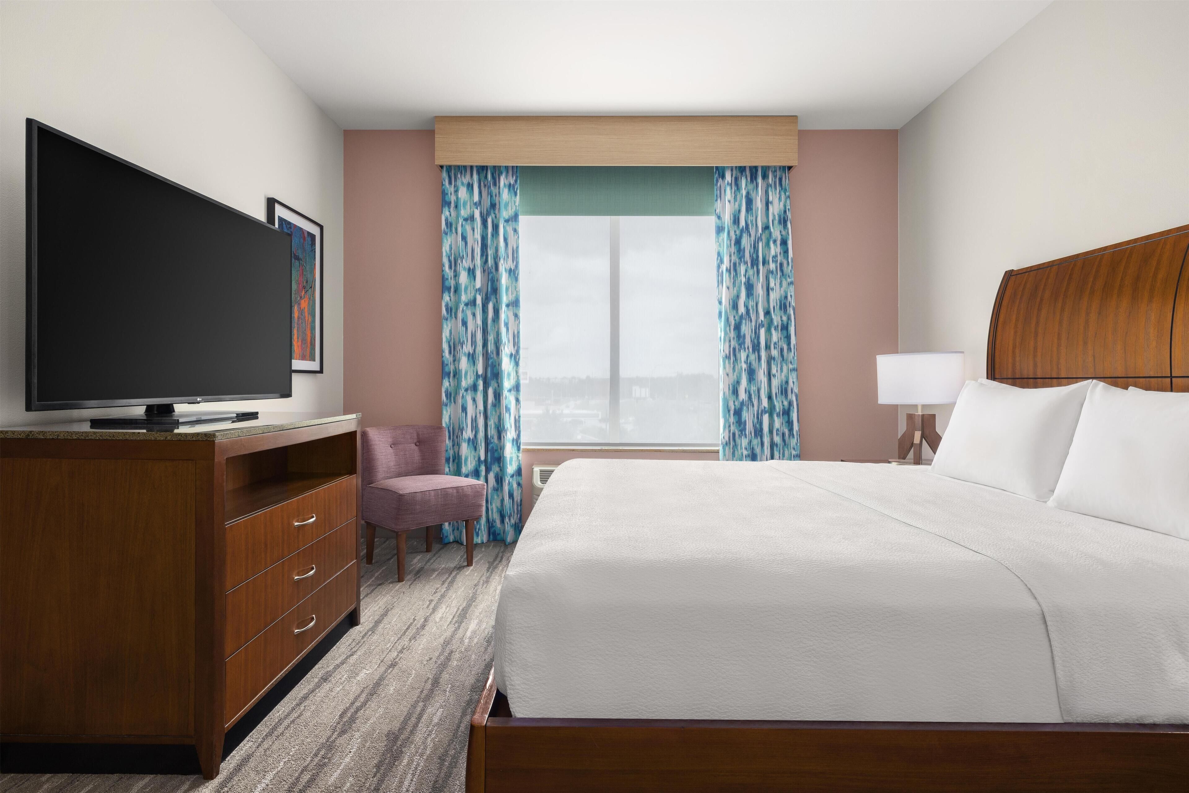 Habitación de hotel con cama, televisión, escritorio, silla y ventana con cortinas.