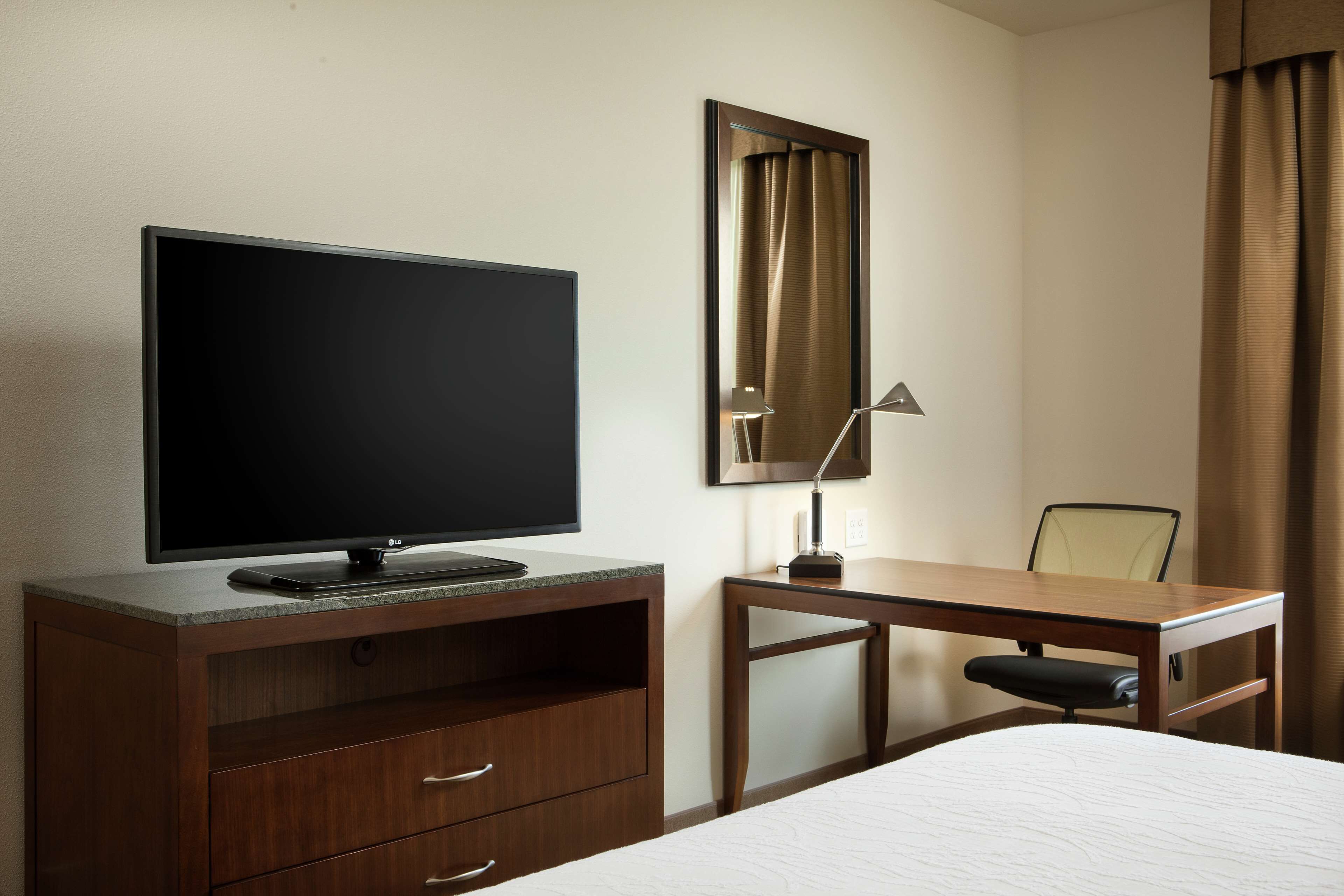 Une chambre d’hôtel équipée d’un téléviseur à écran plat posé sur un meuble en bois, d’un bureau avec une chaise et d’un grand miroir mural.