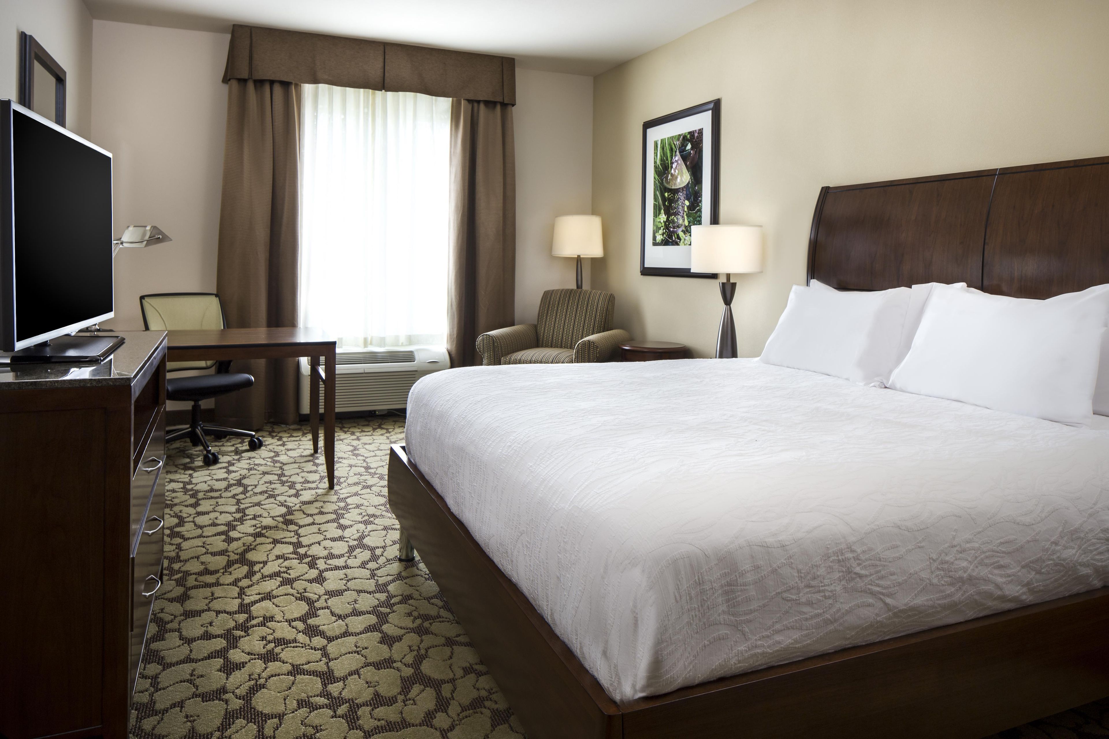 A hotel room with a large bed, a desk with a chair, a television, and a window with curtains.