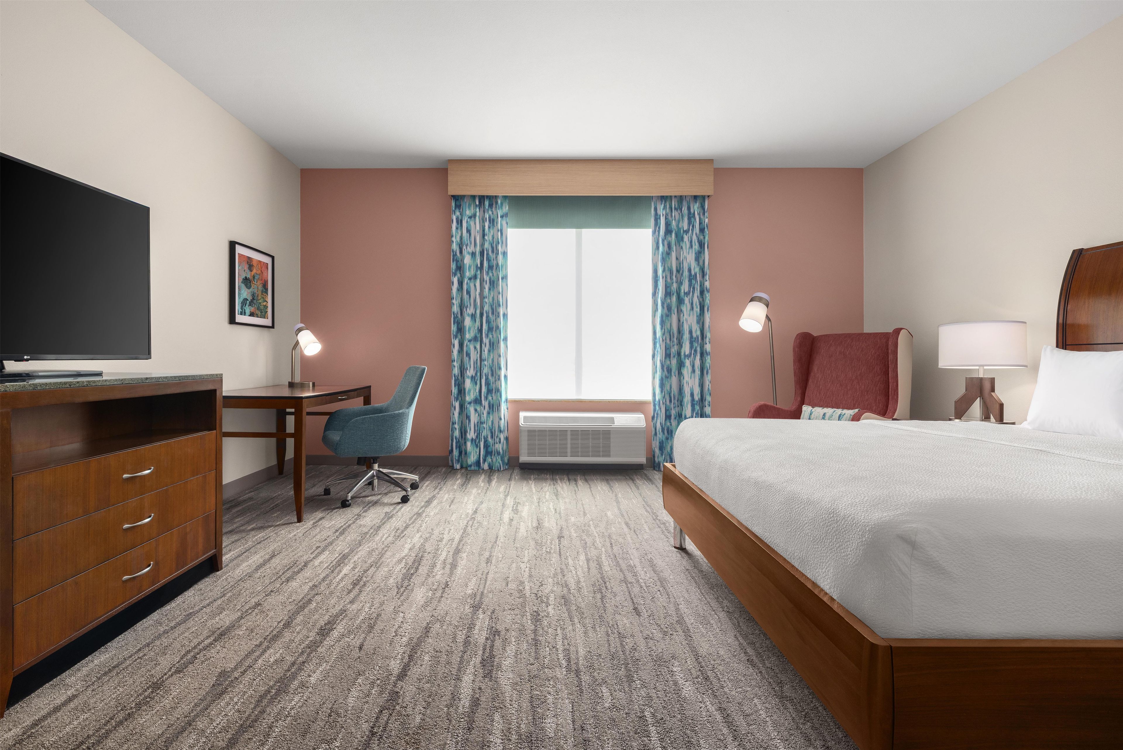 A hotel room with a bed, desk, chair, TV, and a large window with curtains.