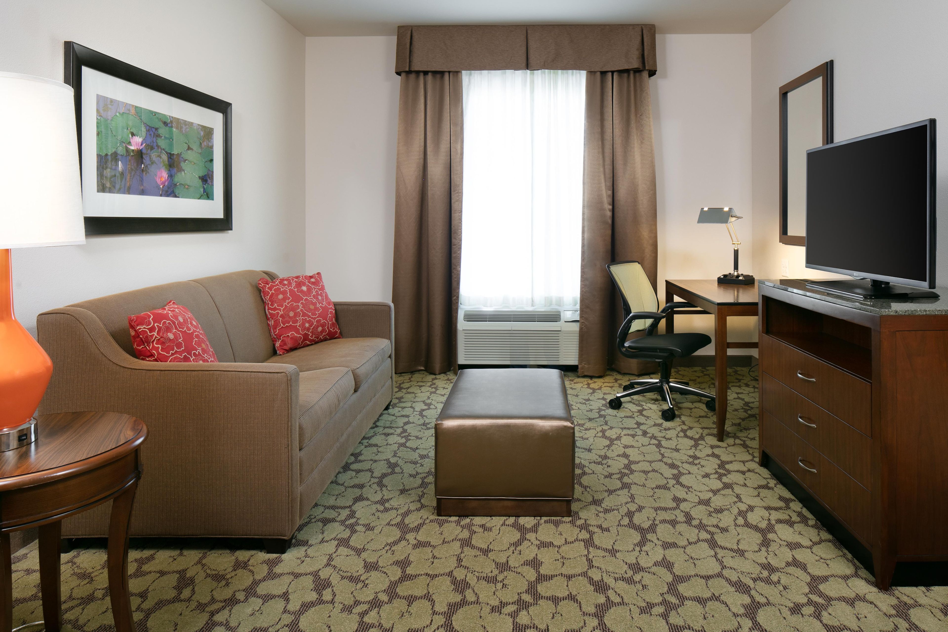 Une chambre d’hôtel équipée d’un canapé, d’un pouf, d’une télévision, d’un bureau, d’une chaise et d’un tableau encadré au mur.