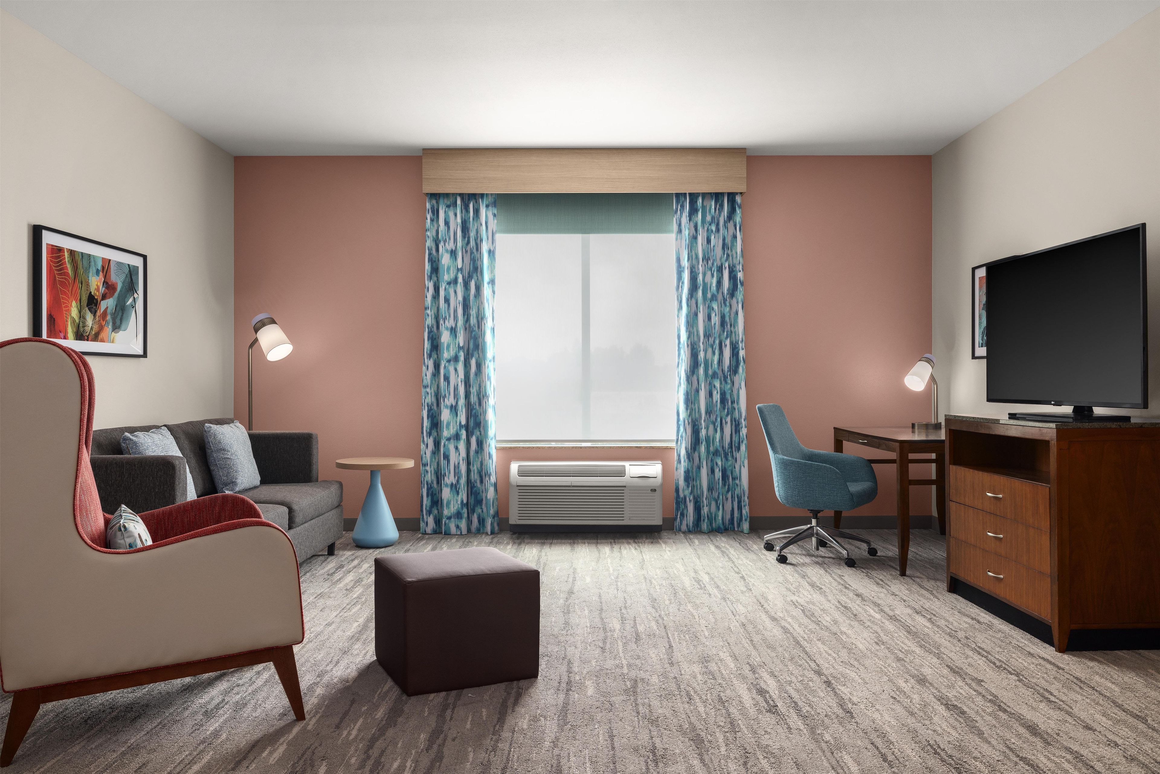 A hotel room with a sofa, ottoman, desk, chair, television, air conditioning unit, and curtains.