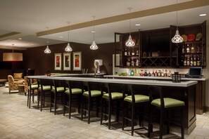 Zona de bar con una barra alargada, varios taburetes, lámparas colgantes y una estantería para vinos con botellas.
