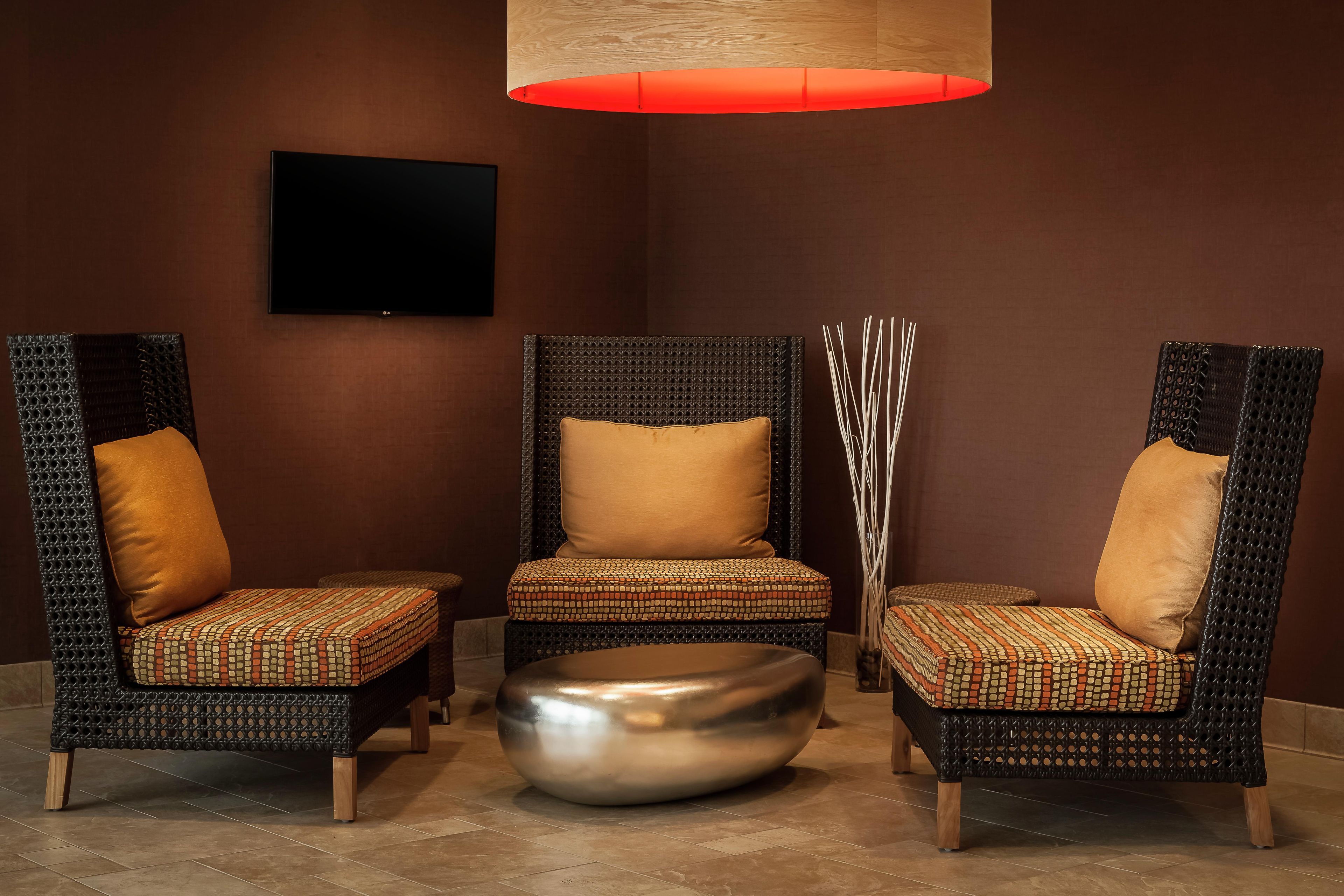 A seating area with three chairs and a central ottoman, all with patterned upholstery, against a brown wall with a flat-screen TV mounted above.