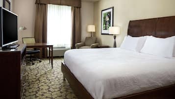 Ein Hotelzimmer mit einem großen Bett, einem Schreibtisch mit Fernseher, einem Sessel und einem Fenster mit Vorhängen.