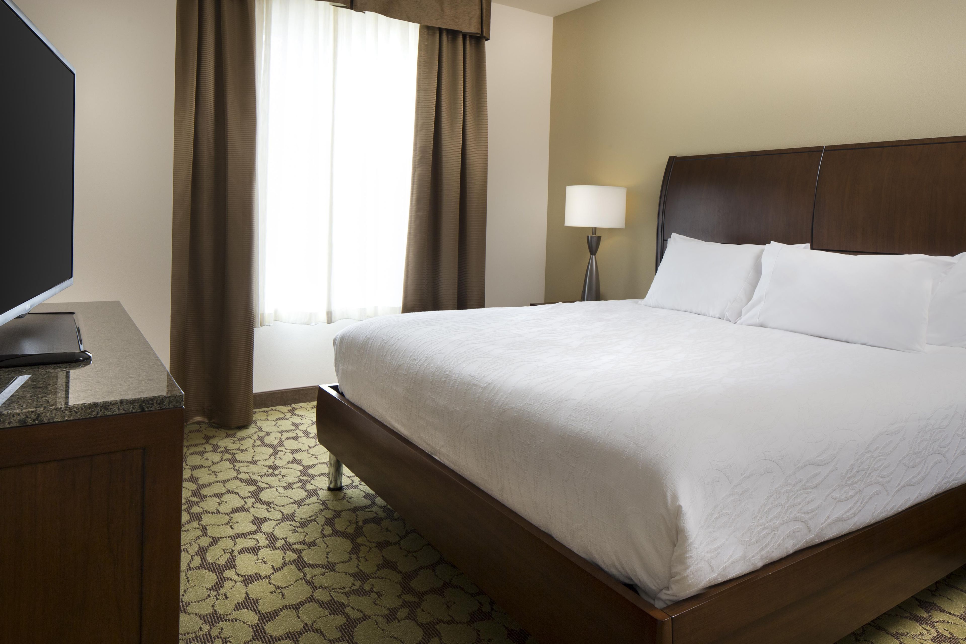 Una habitación de hotel con cama, escritorio, lámpara y ventana con cortinas.