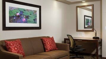 Quarto de hotel com sofá marrom, almofadas estampadas em vermelho, escrivaninha com cadeira, quadro com nenúfares e um espelho.