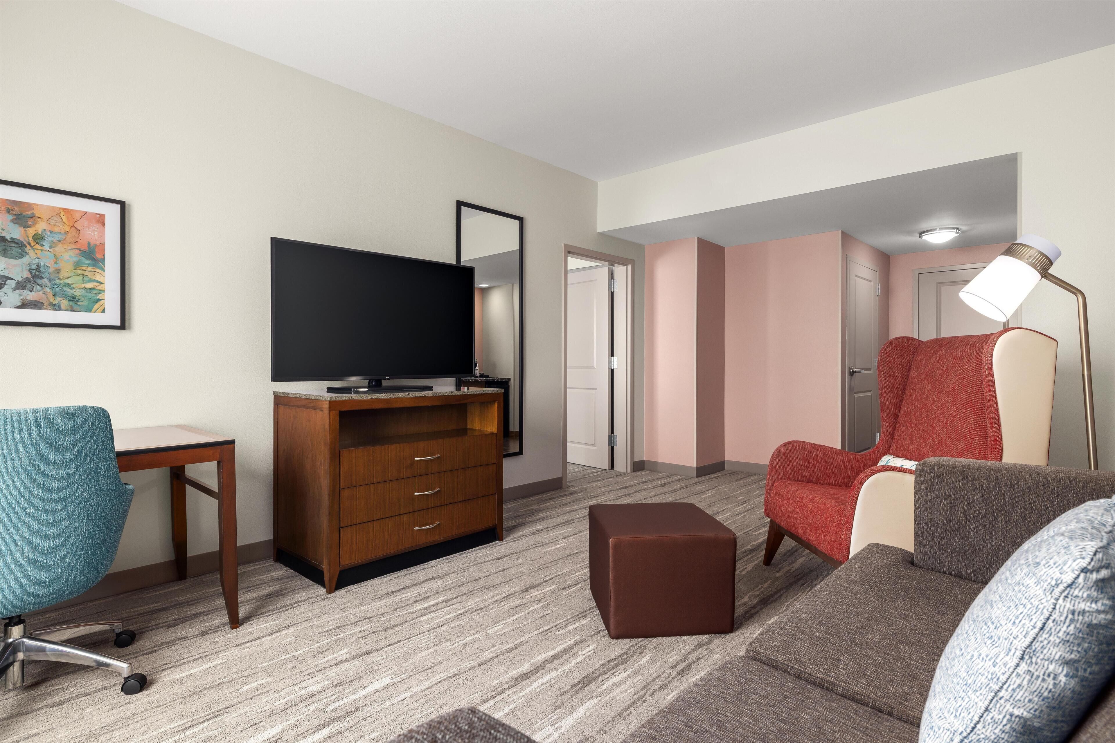 A hotel room with a flat-screen TV, a wooden cabinet, a red armchair, a blue office chair, a brown ottoman, and a floor lamp.
