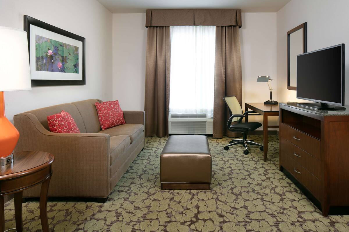 A hotel room with a brown sofa, red pillows, a wooden side table, a brown ottoman, a desk with a chair, a television, and a framed picture on the wall.