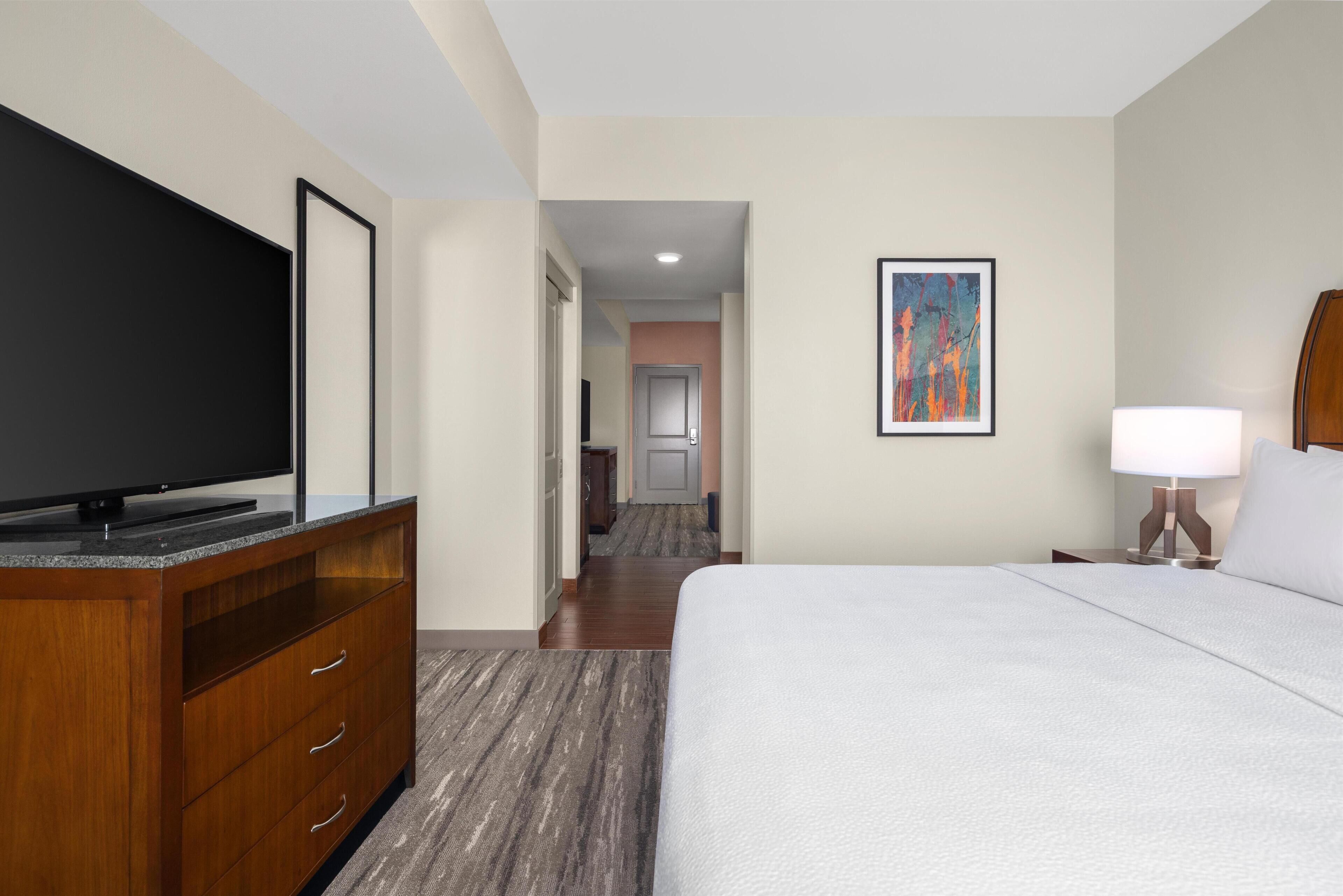 Habitación de hotel con cama, televisor en una cómoda de madera, un cuadro en la pared y un pasillo que conduce a un baño.