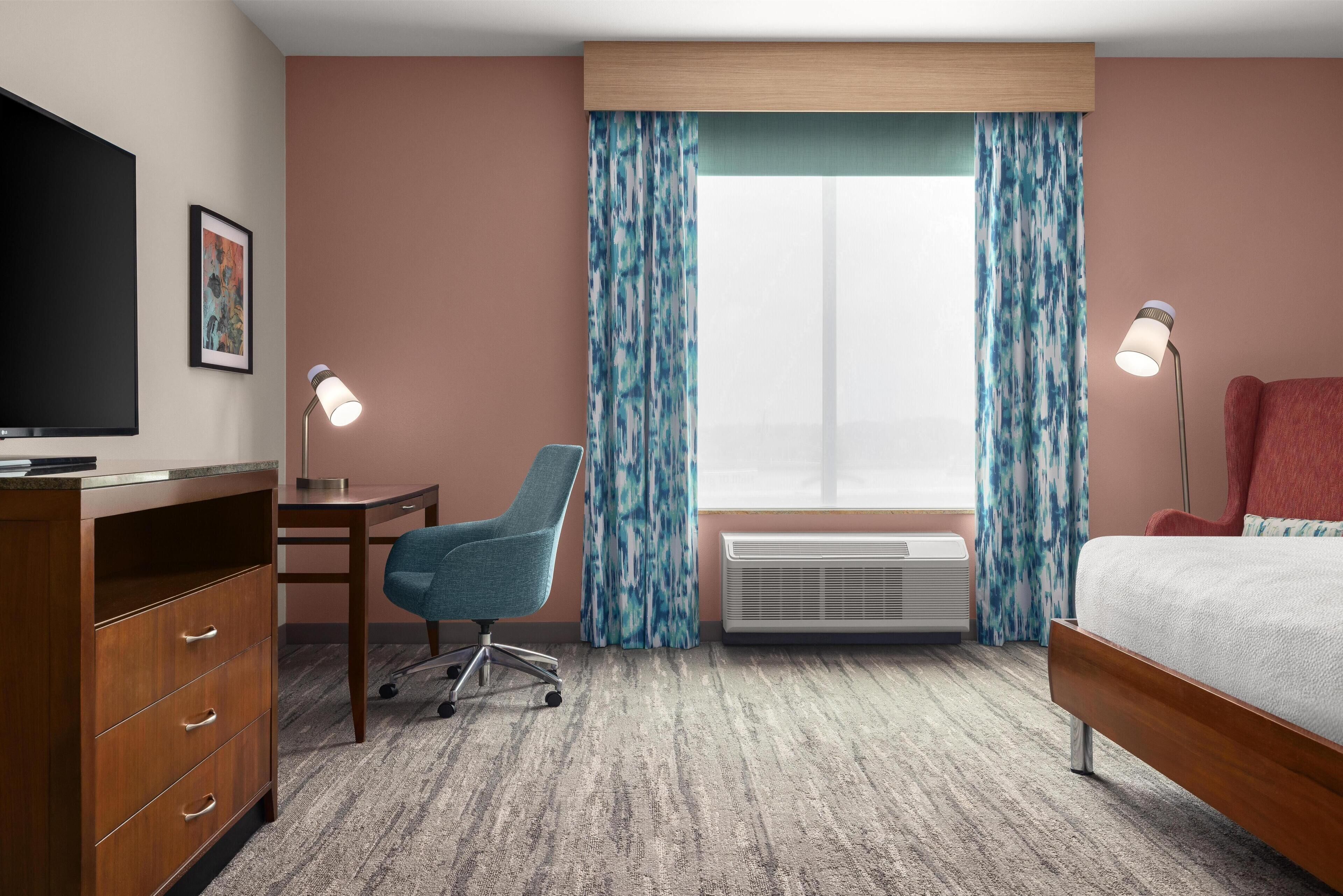 A hotel room with a bed, desk, chair, television, air conditioning unit, and curtains.