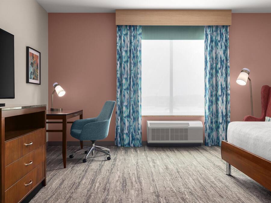 A hotel room with a bed, desk, chair, television, air conditioning unit, and curtains.
