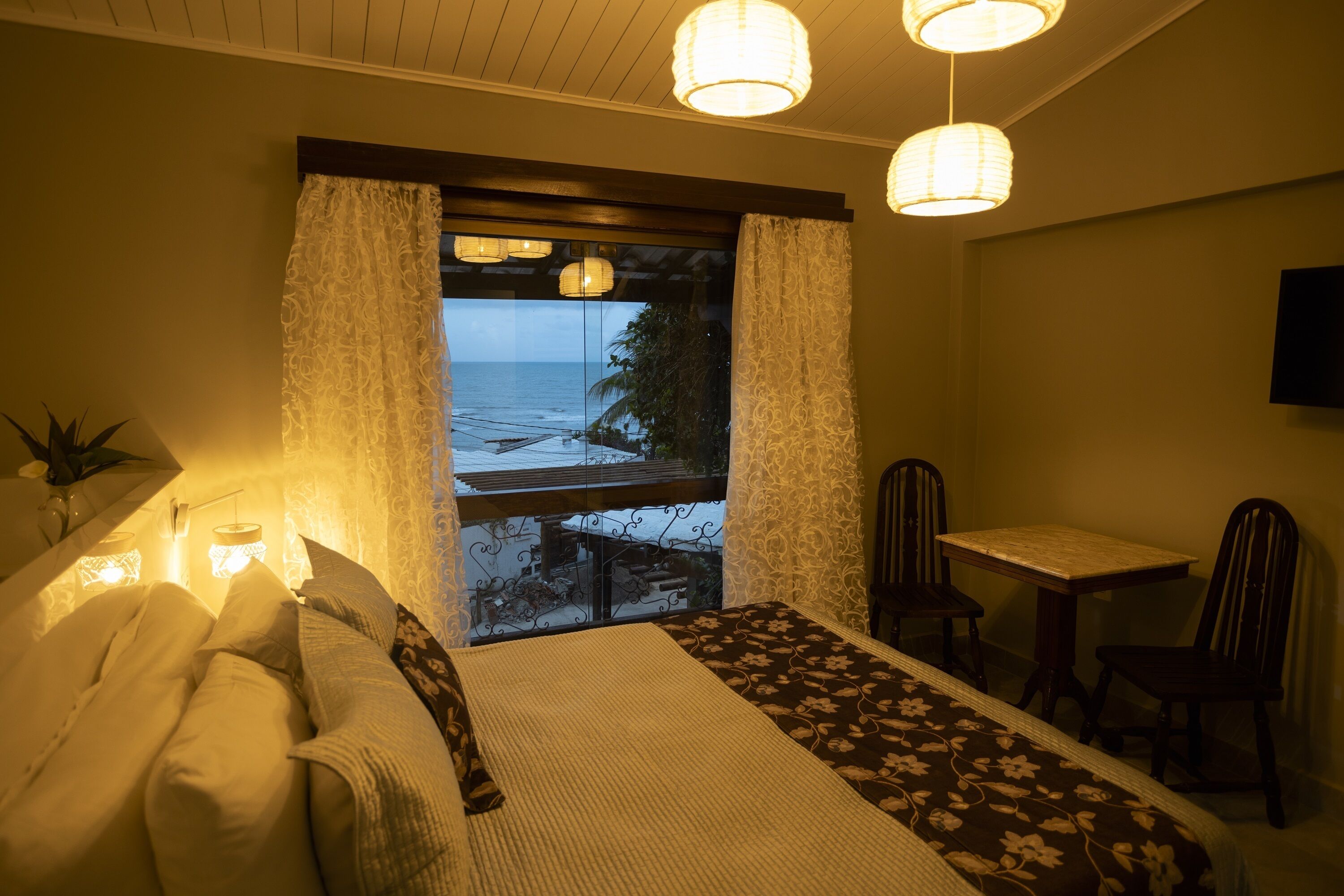 Un dormitorio con cama, una mesita, dos sillas y vista al océano a través de una ventana con cortinas.