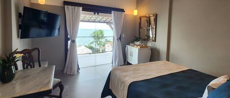 A hotel room with a bed, a desk, a TV, a small cabinet, and a view of the ocean.