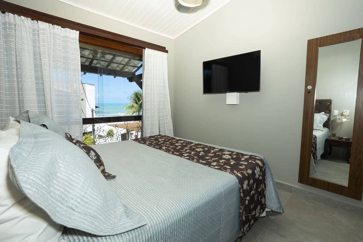 Une chambre à coucher avec un lit, une télévision, un grand miroir et une fenêtre donnant sur l’océan.