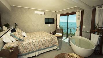 A bedroom with a large bed, a bathtub, a television, and a view of the sea.