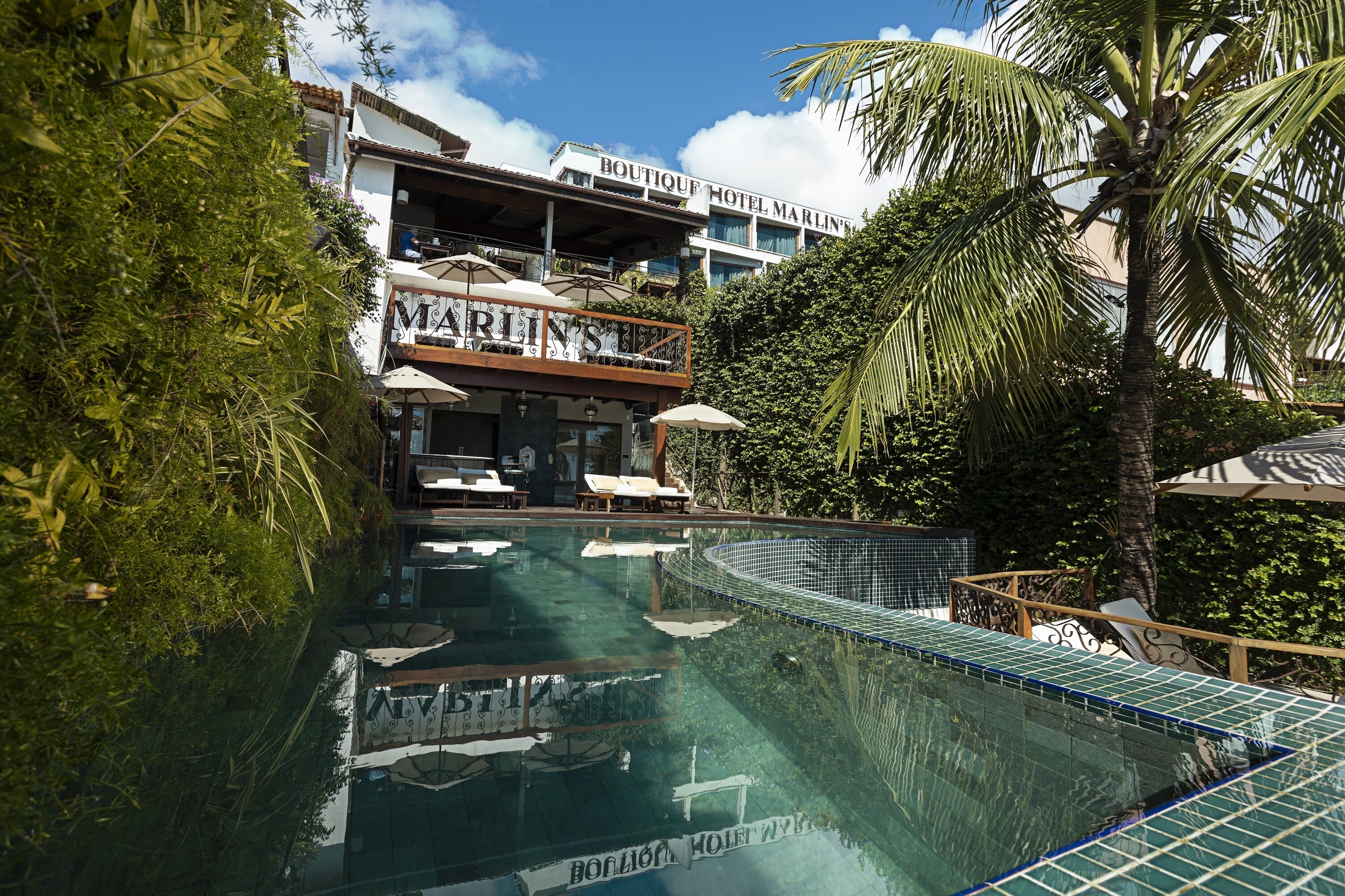 Piscina con sillas de descanso y sombrillas, rodeada de exuberante vegetación y un edificio de hotel con balcones.