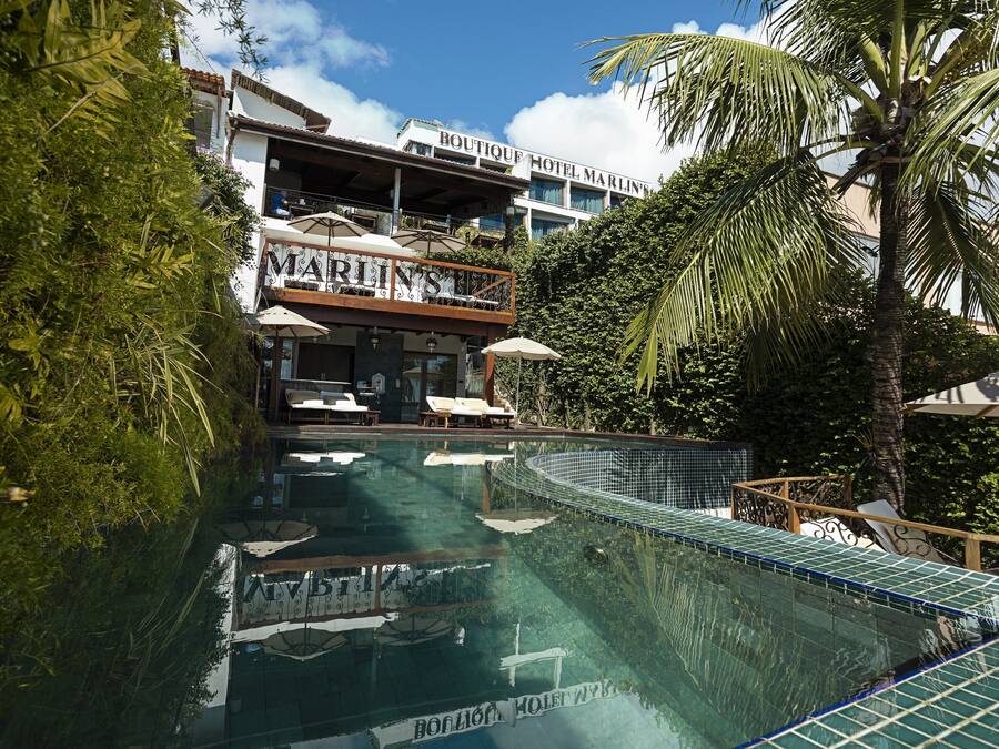 A swimming pool with lounge chairs and umbrellas, surrounded by lush greenery and a hotel building with balconies.