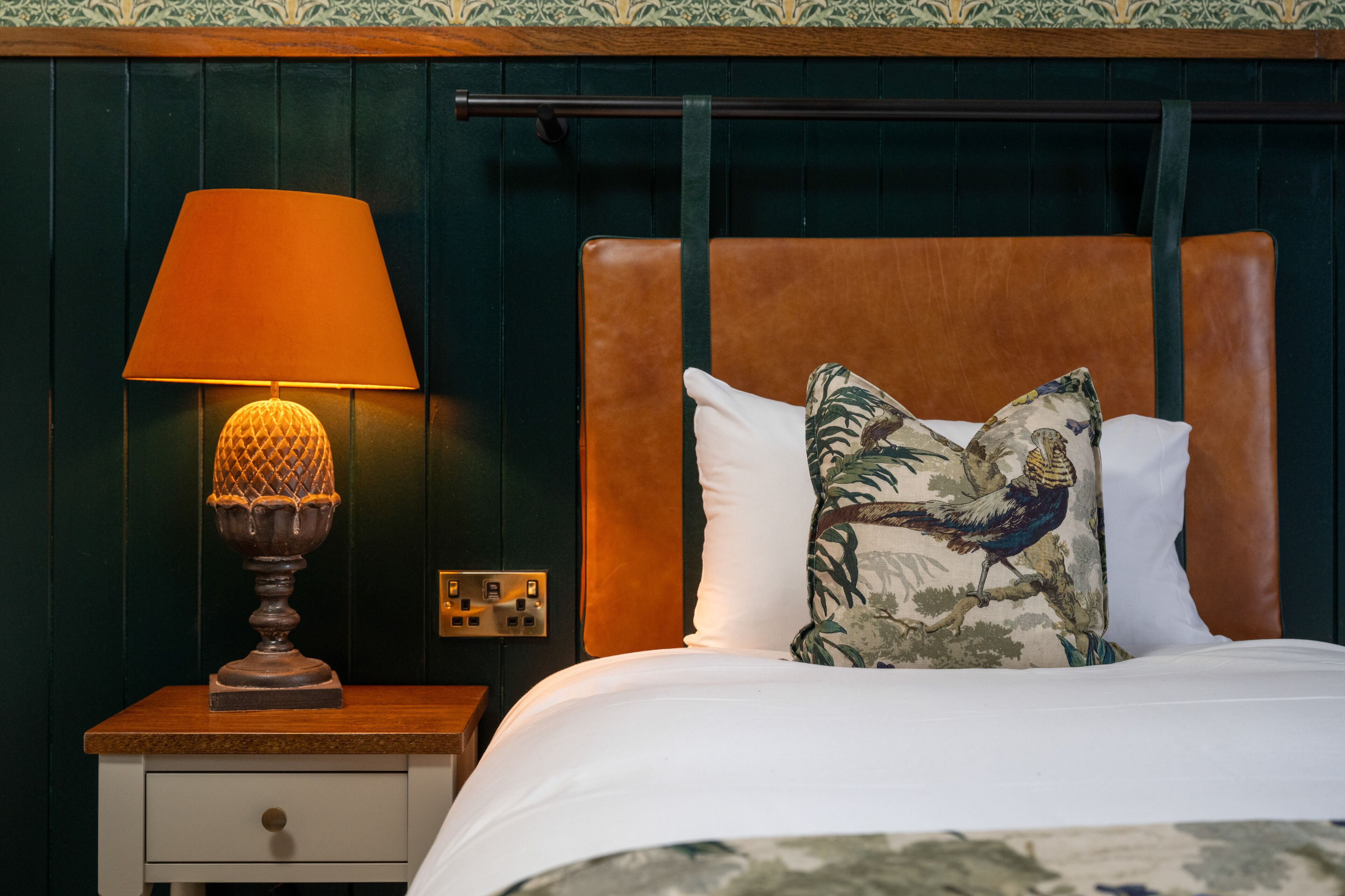 Una cama bien tendida con un cabecero de madera y un cojín decorativo. Una mesita de noche de madera con una lámpara está al lado de la cama.