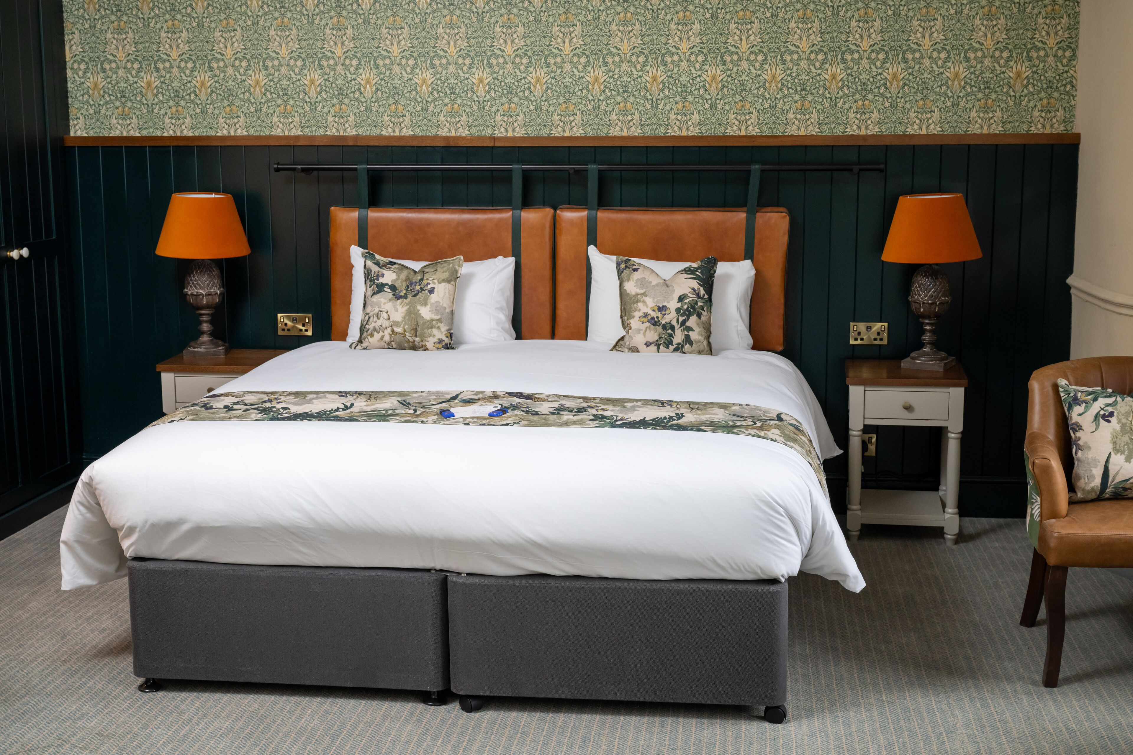 A neatly made bed with a white duvet, two matching pillows, and a floral throw. There are two bedside tables with lamps, a brown leather armchair, and a patterned wallpaper above the bed.
