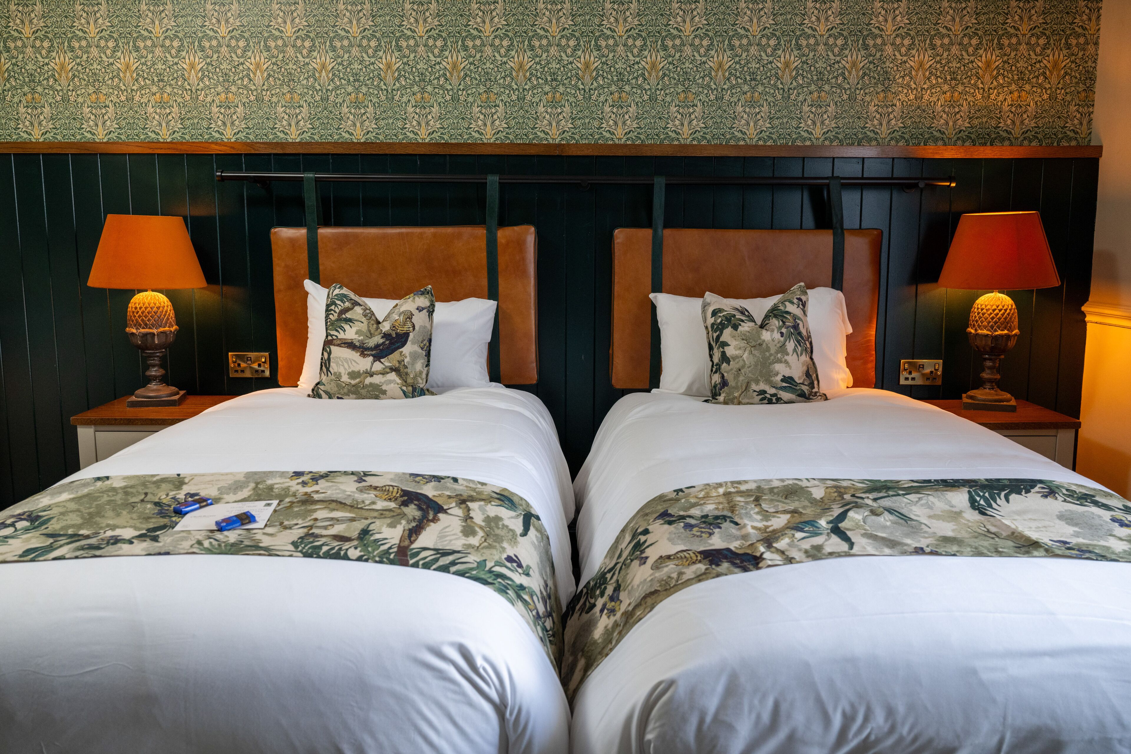 Habitación doble con dos camas, cada una con cabecera, mesita de noche, lámpara y colcha estampada.