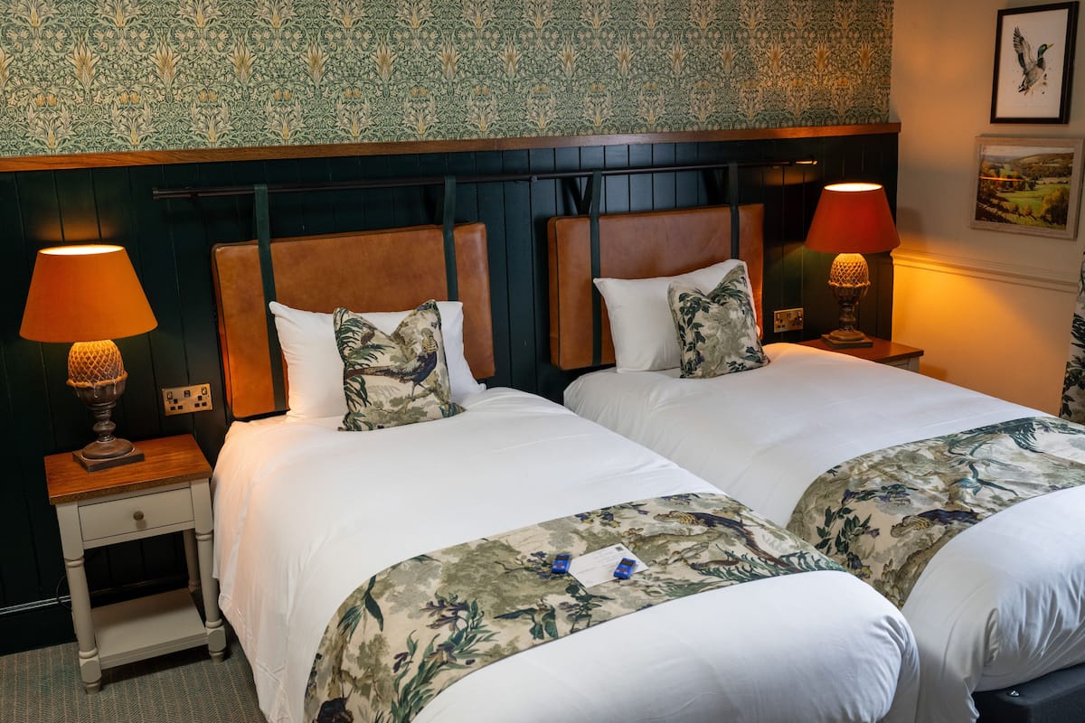 Habitación de hotel con dos camas, cada una con cabecero de madera, ropa de cama blanca y mantas decorativas con estampado floral. Hay dos lámparas de noche con abat-jour rojo y un cuadro en la pared.