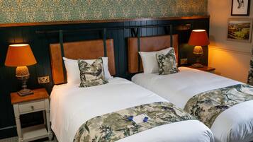 A hotel room with two beds, each with a wooden headboard, white bedding, and floral-patterned throw blankets. There are two bedside lamps with red shades and a framed picture on the wall.