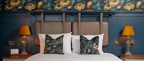 A neatly made bed with white linens and floral-patterned pillows, flanked by two bedside tables with lamps, set against a wall with a floral wallpaper.