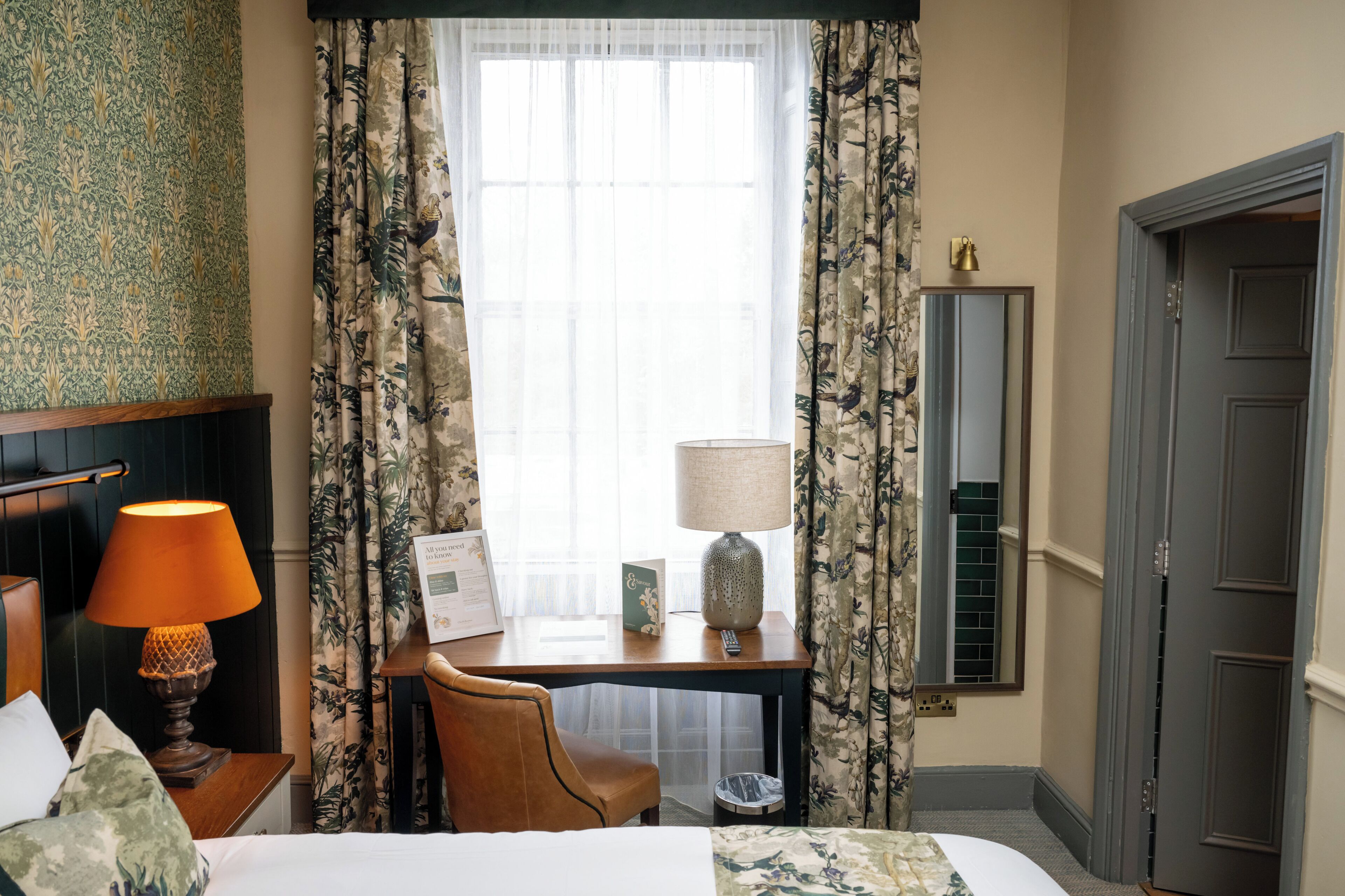 Habitación de hotel con cama, escritorio con lámpara, silla y ventana con cortinas.