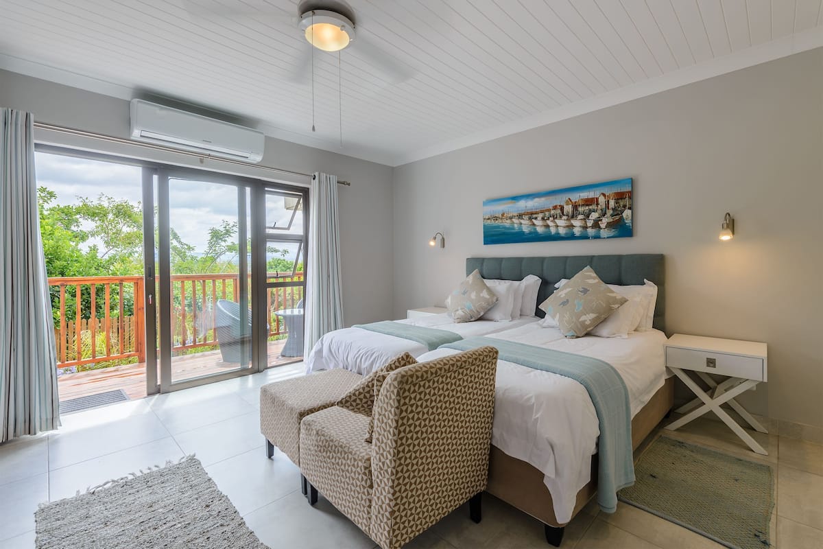 A bedroom with a bed, two bedside tables, a chair, a painting, and a view through a glass door to an outdoor area.