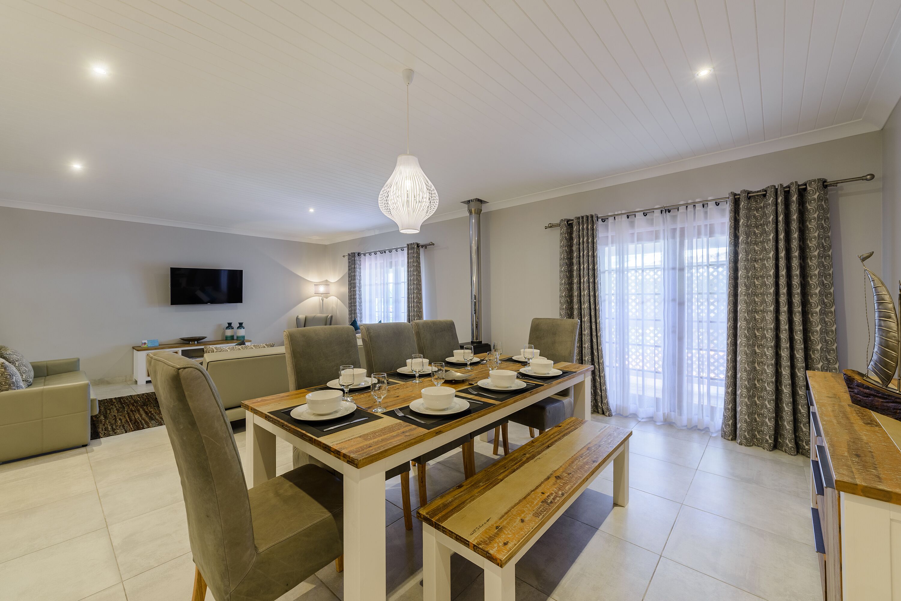 Un comedor con una mesa de madera dispuesta para seis personas, rodeada de sillas, un banco y un televisor montado en la pared.