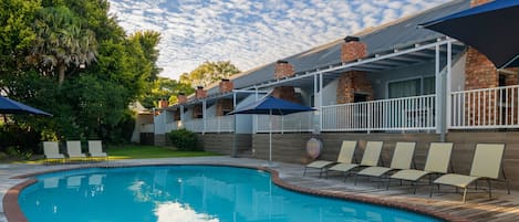 Una piscina con borde de ladrillo, rodeada de sillones de descanso y sombrillas, y un área de patio techada con muros y techo de ladrillo.