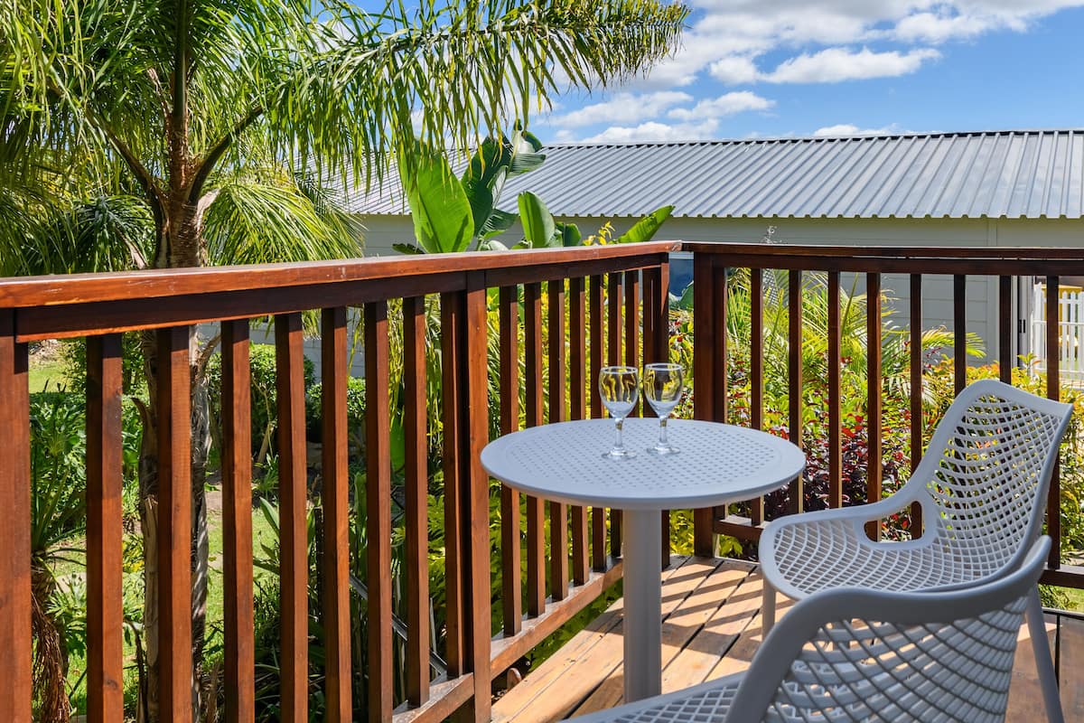 A wooden deck with a round table and two chairs, surrounded by lush greenery and a building with a corrugated metal roof.
